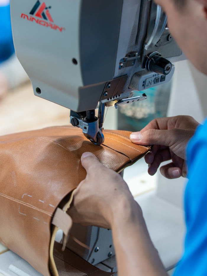 A machinist guiding a tan leather upper under the needle of a post-bed sewing machine.
