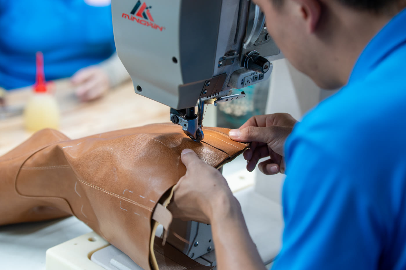Two hands guiding a tan leather upper under the needle of a closing machine.
