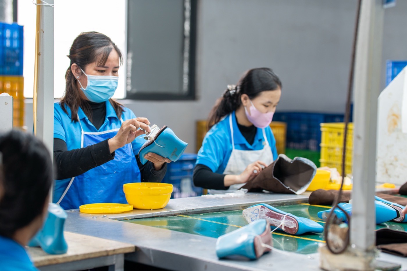 Two workers at the cementing bench, one holding a pale blue heeled upper, sole units and lasts on the table between them.