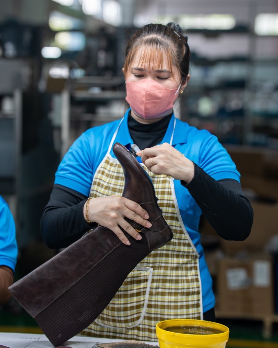 A worker in an apron dressing the shaft of a dark brown leather boot by hand.