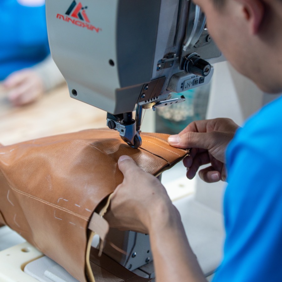 Hands guiding a tan leather upper under the needle of a post-bed stitching machine.