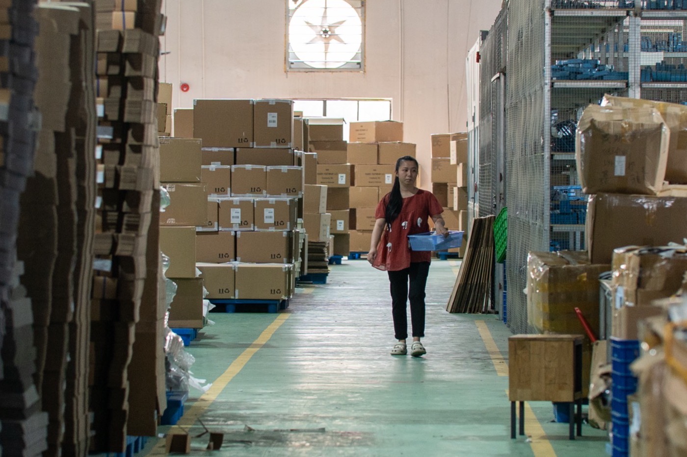 A worker walking an aisle of the finished-goods store between stacked cartons.
