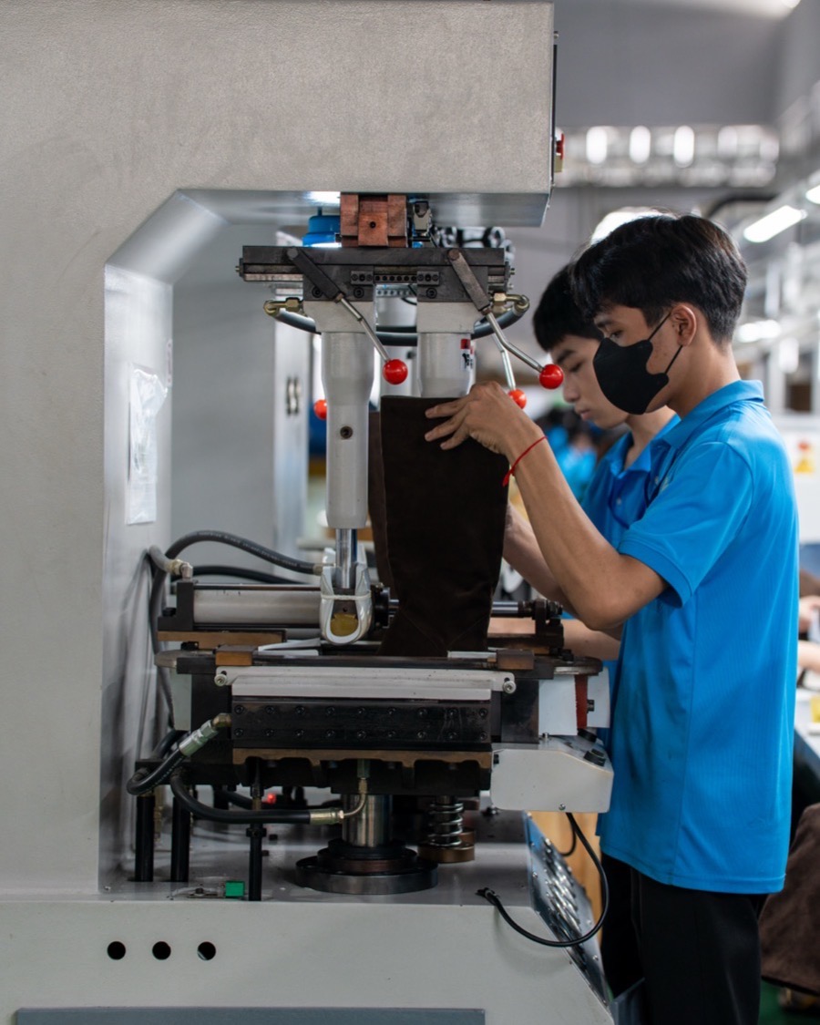 A worker at a hydraulic press, shaping a dark suede upper between the plates.